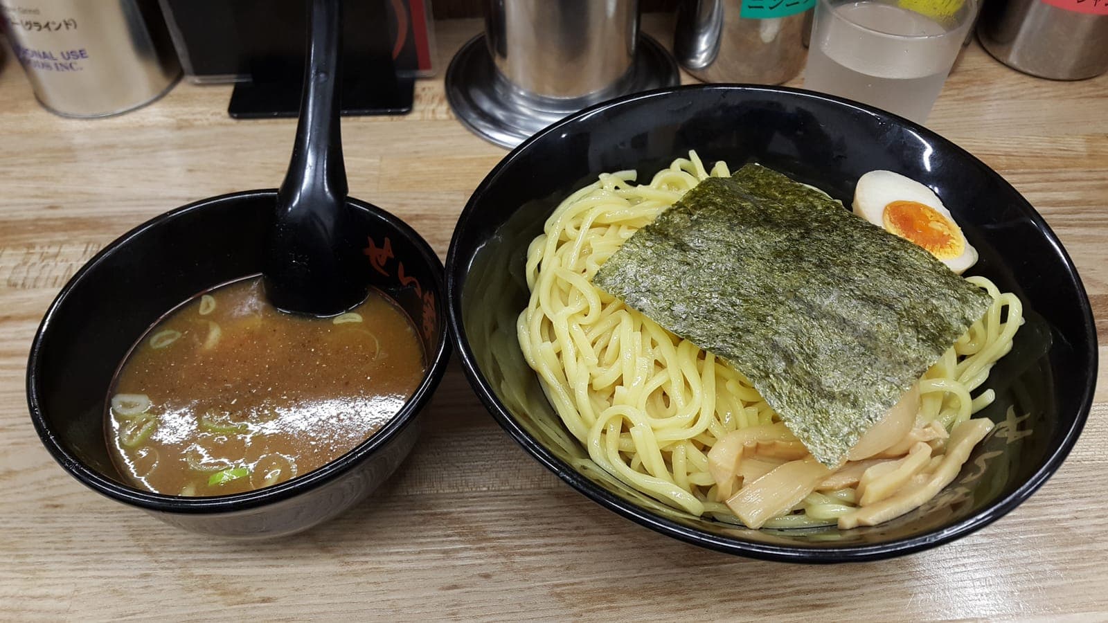 A bowl of yellow wheat noodles with nori and a soft egg beside a bowl of dipping broth