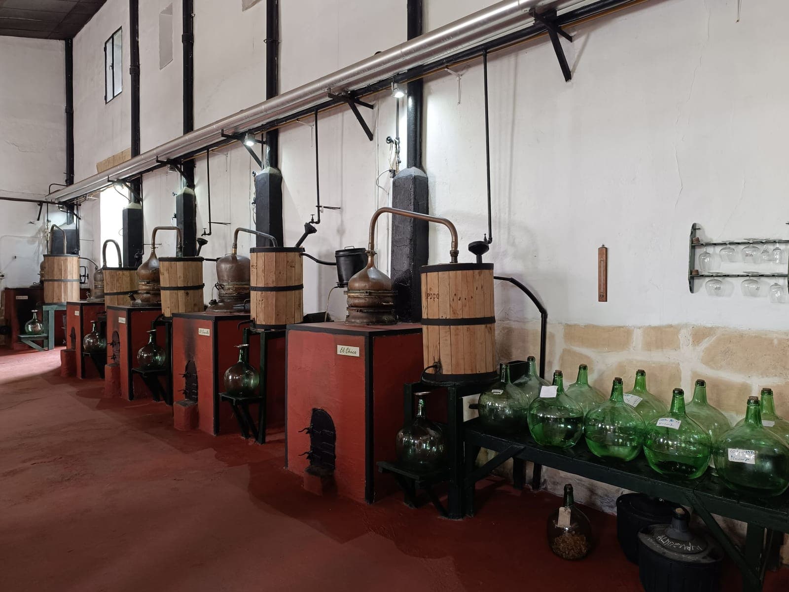 A row of small copper pot stills along a distillery wall with glass demijohns below