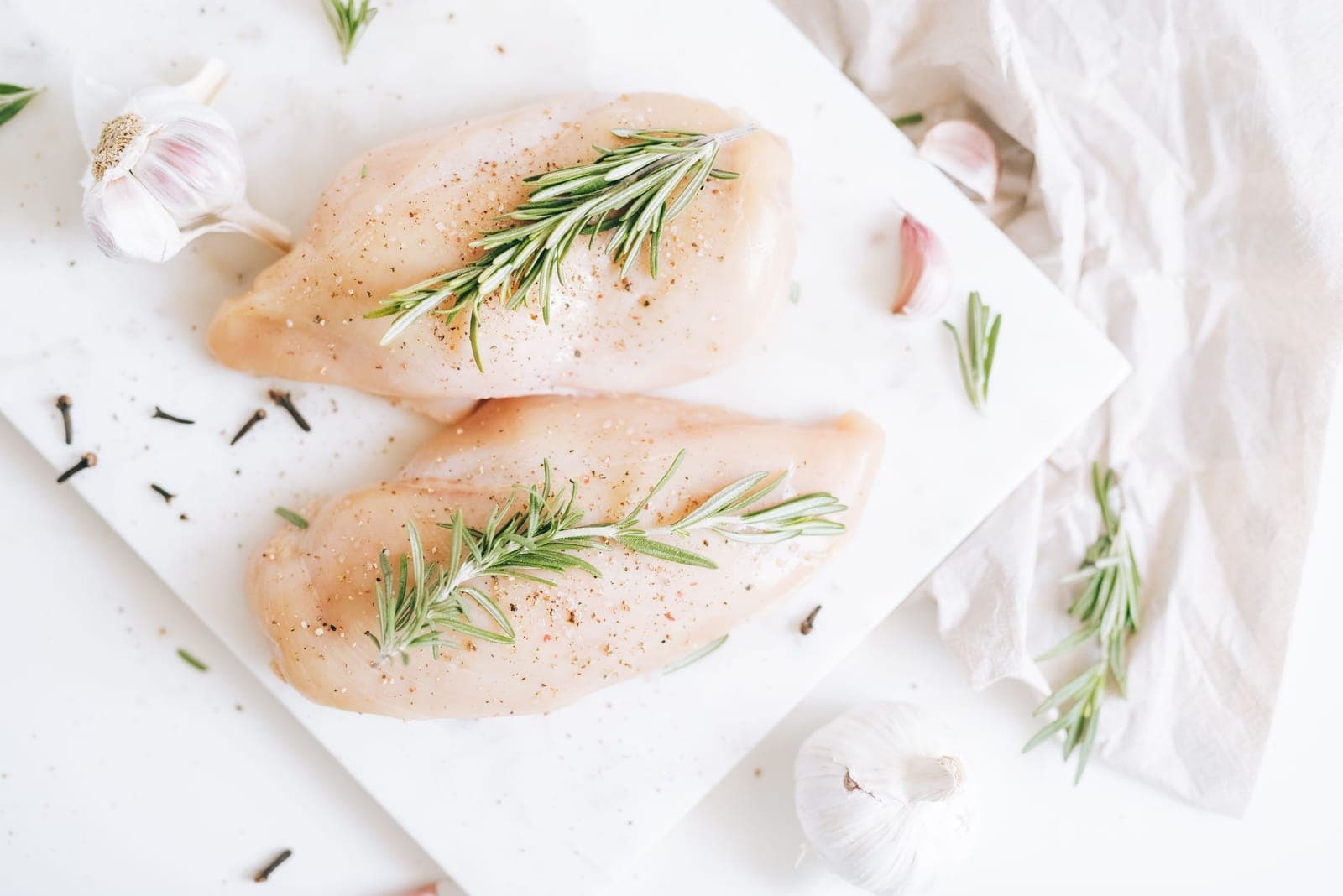 Two raw chicken breasts seasoned with salt, pepper and rosemary on a marble board