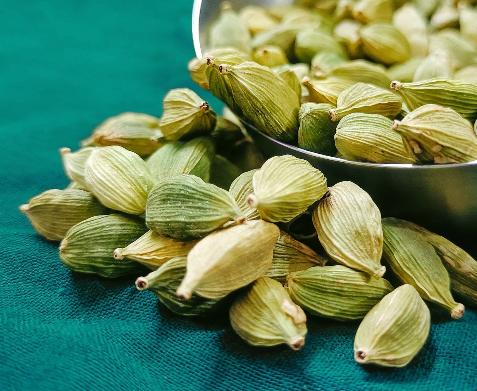 Green cardamom pods spilling from a metal dish onto teal cloth