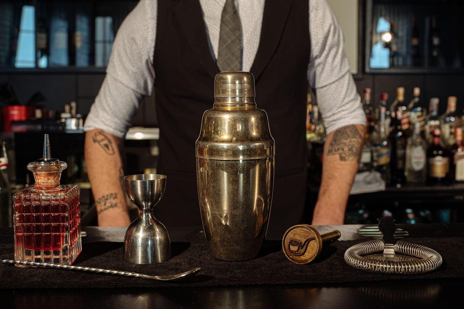 A cocktail shaker, jigger and strainer set out on a bar top in front of a bartender