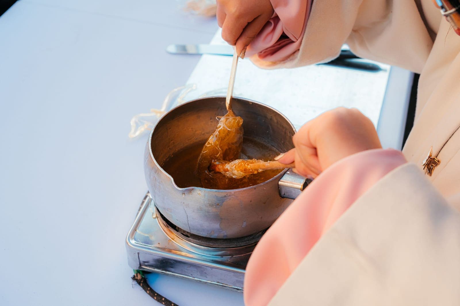 Hands pulling a thread of hot sugar syrup from a small pan on a burner