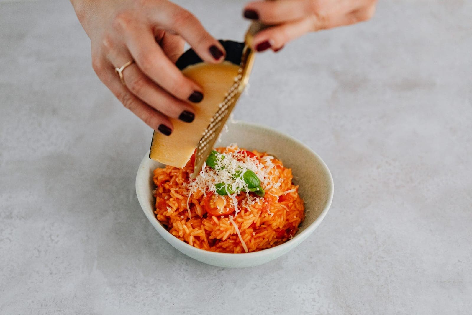 A hand grating hard cheese over a bowl of tomato rice with basil